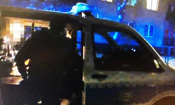 Policjant stoi przy radiowozie tyłem do aparatu i pomaga mężczyźnie