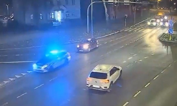 Widok z kamery przemysłowej na przejazd radiowozu, za którym jedzie inny samochód. W głębi widoczne inne samochody jadące ulicą.