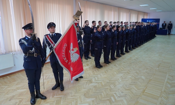 Policjanci stoją w dwuszeregu podczas uroczystości ślubowania. Na pierwszym planie trzej policjanci z pocztu sztandarowego, środkowy z nich trzyma sztandar, pozostali dwaj trzymają szable uniesione do góry.