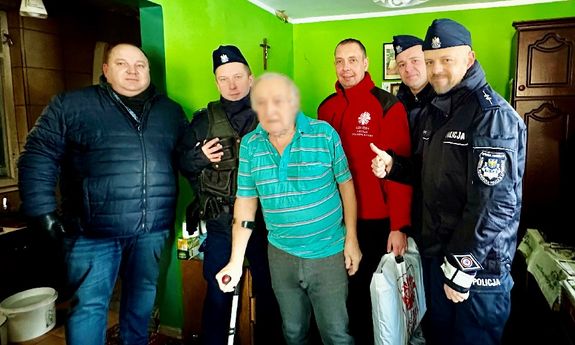 W pokoju z zielonymi ścianami stoją policjanci i ksiądz. Przed nimi stoi mężczyzna, któremu pomagają darami