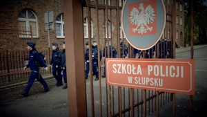 grupa policjantów maszeruje, na pierwszym planie tabliczka na bramie z napisem Szkoła Policji w Słupsku, a nad nim godło Polski