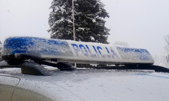 Dach radiowozu oznakowanego. Na dachu belka z napisem Policja. Samochód przysypany śniegiem. W tle zaśnieżone drzewo iglaste. Sceneria zimowa