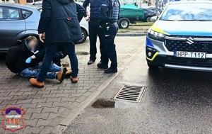 Radiowóz  oraz siedzący na ziemi mężczyzna. Obok niego umundurowani policjanci i mężczyzna.