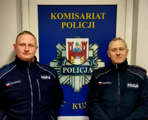 dwóch umundurowanych policjantów stoi na tle baneru Komisariatu Policji