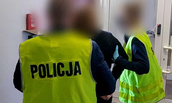 Dwaj policjanci w żółtych kamizelkach z napisem policja prowadza zatrzymanego mężczyznę.