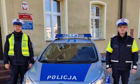 Dwóch policjantów stoi przy oznakowanym radiowozie. W tle budynek policji