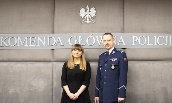Komendant Główny Policji stoi z Rzeczniczką Praw dziecka na szarym tle. Na ścianie napis Komenda Główna Policji