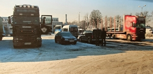 Zaparkowany samochód nieopodal znajdujących się tam aut ciężarowych, obok niego stoją dwaj mężczyźni