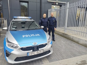Dwaj policjanci stoją przy radiowozie na parkingu przed budynkiem.