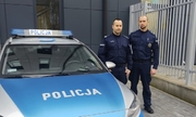 Dwaj policjanci stoją przy radiowozie na parkingu przed budynkiem.