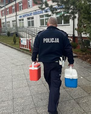 Idący w kierunku Szpitalnego Oddziału Ratunkowego umundurowany policjant ruchu drogowego. W prawej ręce trzyma pojemnik koloru niebieskiego, w lewej czerwonego