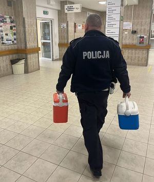 Idący w kierunku Szpitalnego Oddziału Ratunkowego umundurowany policjant ruchu drogowego. W prawej ręce trzyma pojemnik koloru niebieskiego, w lewej czerwonego