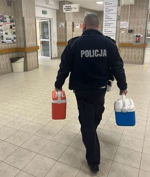 Idący w kierunku Szpitalnego Oddziału Ratunkowego umundurowany policjant ruchu drogowego. W prawej ręce trzyma pojemnik koloru niebieskiego, w lewej czerwonego