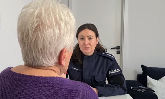 Na pierwszym planie plecy kobiety. W tle siedzi policjantka