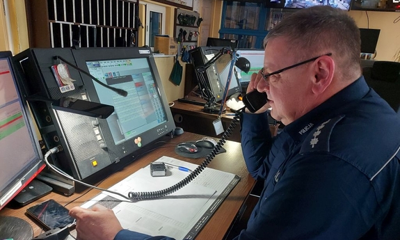 Dyżurny policji, policjant siedzi przy biurku na stanowisku kierowania jednostką. Rozmawia przez telefon. Przed nim monitory komputerów, dalej wnętrze pomieszczenia służbowego