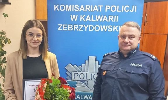 aNa zdjęciu po lewej stronie kobieta w stroju cywilnym trzymająca w ręku kwiaty i list gratulacyjny, po prawej umundurowany policjant. W tle policyjny baner z napisem Komisariat Policji w Kalwarii Zebrzydowskiej