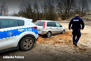 Policjant idzie koło dwóch samochodów. Jeden to radiowóz, drugi to samochód, który ugrzązł w błocie