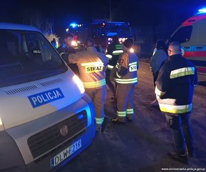 Pora nocna. Funkcjonariusze Policji i Straży Pożarnej stoją przy policyjnym radiowozie. Z boku widać karetkę pogotowia i wóz straży pożarnej