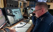 Policjant na stanowisku kierowania jednostką rozmawia przez telefon. Zdjęcie zrobione w pomieszczeniu dla dyżurnych policji, w tle komputery i monitory.