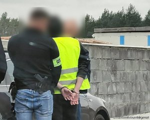 policjant zakłada kajdanki zatrzymanemu mężczyźnie. Obaj stoją przy samochodzie. Widok z tyłu