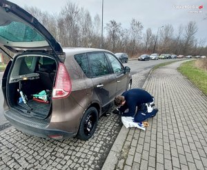 Zdjęcie przedstawia umundurowanego policjanta wymieniającego koło w samochodzie. W tle samochody oraz jezdnia