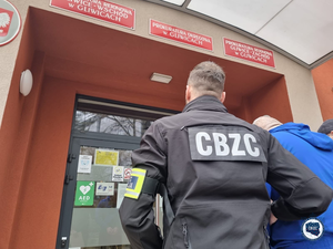 policjant ma na kurtce napis CBZC. Prowadzi do budynku zatrzymanego mężczyznę