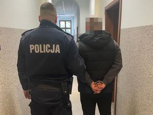 policjant w polarze z napisem Policja oraz zatrzymanego mężczyznę w kajdankach założonych na ręce trzymane z tyłu