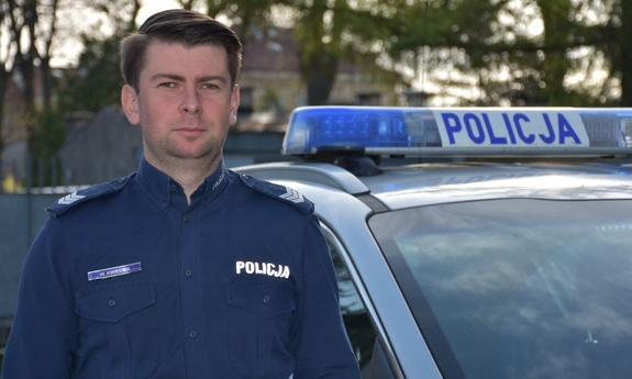 na zdjęciu umundurowany policjant, w tle radiowóz policyjny