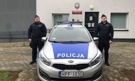 dwóch umundurowanych policjantów stoi przy radiowozie, w tle budynek jednostki policji