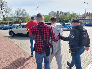 dwaj ubrani po cywilnemu policjanci prowadzą zatrzymanego. Przed nimi idzie trzeci policjant