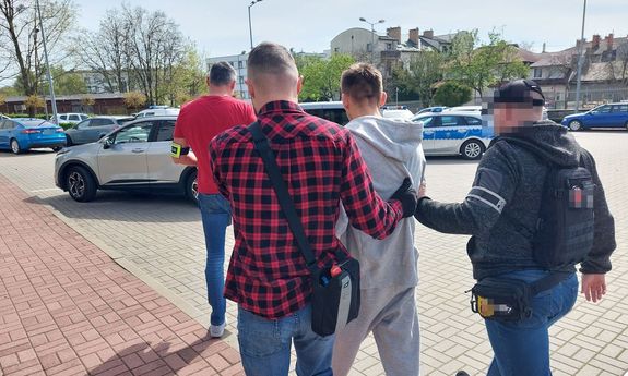 dwaj ubrani po cywilnemu policjanci prowadzą zatrzymanego. Przed nimi idzie trzeci policjant