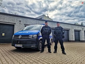 Zdjęcie przedstawia dwóch umundurowanych policjantów na tle oznakowanego policyjnego radiowozu