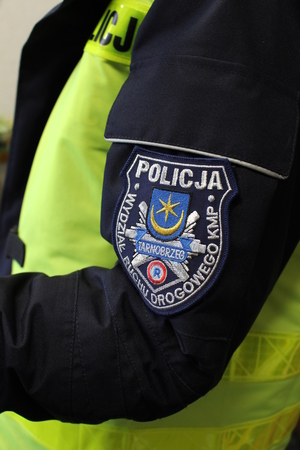 Zdjęcie poglądowe. Fragment funkcjonariusza stojącego bokiem. Na ramieniu munduru naszywka z logo Wydziału Ruchu Drogowego KMP Tarnobrzeg