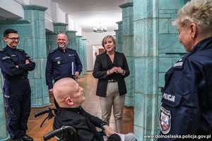 Chłopak na wózku inwalidzkim w holu komendy wojewódzkiej policji wraz z policjantami i pracownicą cywilną