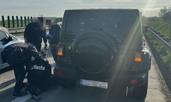 Stojący na autostradzie samochód marki Jeep oraz stojący przy nim funkcjonariusze Policji, w tym mundurowi z ruchu drogowego oraz siedzący na jezdni zatrzymany mężczyzna