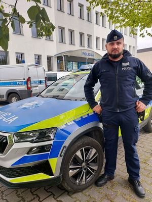 policjant stoi przy radiowozie, w tle budynek komendy oraz radiowozy