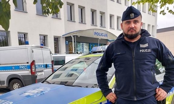 policjant stoi przy radiowozie, w tle budynek komendy oraz radiowozy