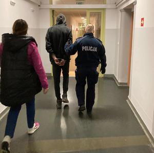 Widok z tyłu. Umundurowany policjant prowadzi zatrzymanego korytarzem. Zatrzymany ma na rękach trzymanych z tyłu założone kajdanki. Za nimi idzie nieumundurowana policjantka.