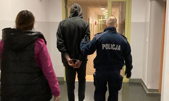 Widok z tyłu. Umundurowany policjant prowadzi zatrzymanego korytarzem. Zatrzymany ma na rękach trzymanych z tyłu założone kajdanki. Za nimi idzie nieumundurowana policjantka.