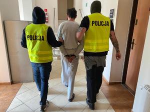 dwaj nieumundurowani policjanci w żółtych kamizelkach z napisem Policja na plecach i w kominiarkach prowadzą zatrzymanego