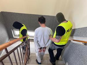 dwaj nieumundurowani policjanci w żółtych kamizelkach z napisem Policja na plecach i w kominiarkach prowadzą zatrzymanego
