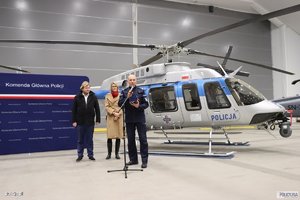 śmigłowiec policyjny w hangarze, przed nim stoi nadinsp. Roman Kuster przemawiający do mikrofonu, za nim stoi kobieta i mężczyzna