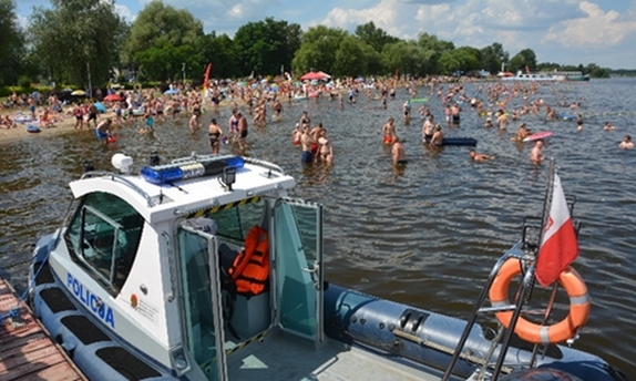na pierwszym planie łódź policyjna na zbiorniku wodnym, w tle ludzie na plaży