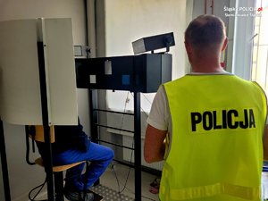 policjant technik stoi obok stanowiska do robienia zdjęć, na którym siedzi zatrzymany