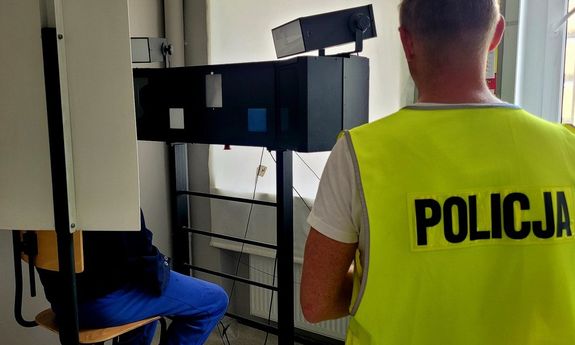 policjant technik stoi obok stanowiska do robienia zdjęć, na którym siedzi zatrzymany