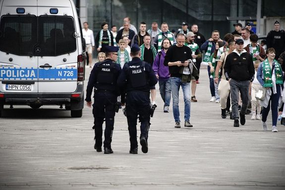 dwaj umundurowani policjanci idą pośród tłumu ludzi. Z boku widoczny policyjny radiowóz