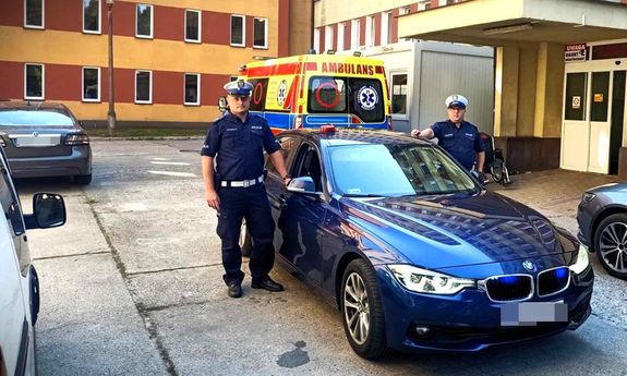 dwaj umundurowani policjanci przy nieoznakowanym radiowozie zaparkowanym przed szpitalem. Z tyłu widać karetkę pogotowia