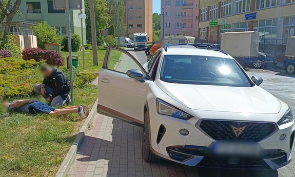zatrzymany leży na trawniku przy ulicy, policjant zakłada mu kajdanki na ręce trzymane za plecami, na chodniku obok stoi samochód osobowy w kolorze białym z otwartymi drzwiami od strony pasażera