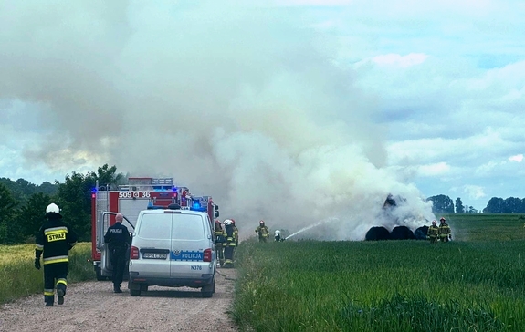 wozy strażackie i radiowóz policyjny na polnej drodze przy polu gdzie płonie sterta balotów ze słomą, która gaszą strażacy, przy radiowozie policjant