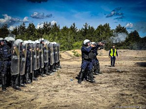 grupa funkcjonariuszy w dwóch rzędach z przodu czterech trzymających gotową do strzału broń gładkolufową oraz Policjanci w drugim rzędzie idący zwartym szykiem , tak zwaną tyralierą